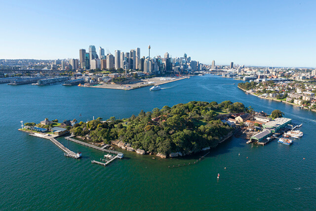 A view of Me-Mel from above showing the island surrounded by the blue water of Sydney Harbour and the Sydney city scape behind it.
