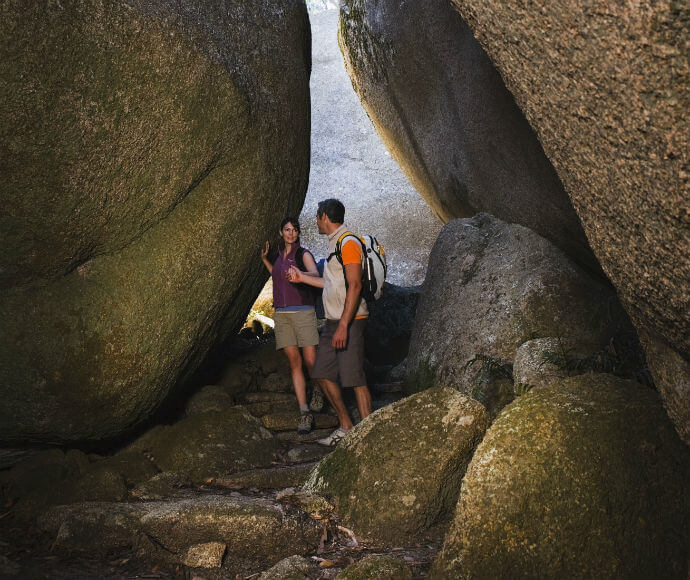Two bushwalkers standing on a rocky path between 2 large boulders.