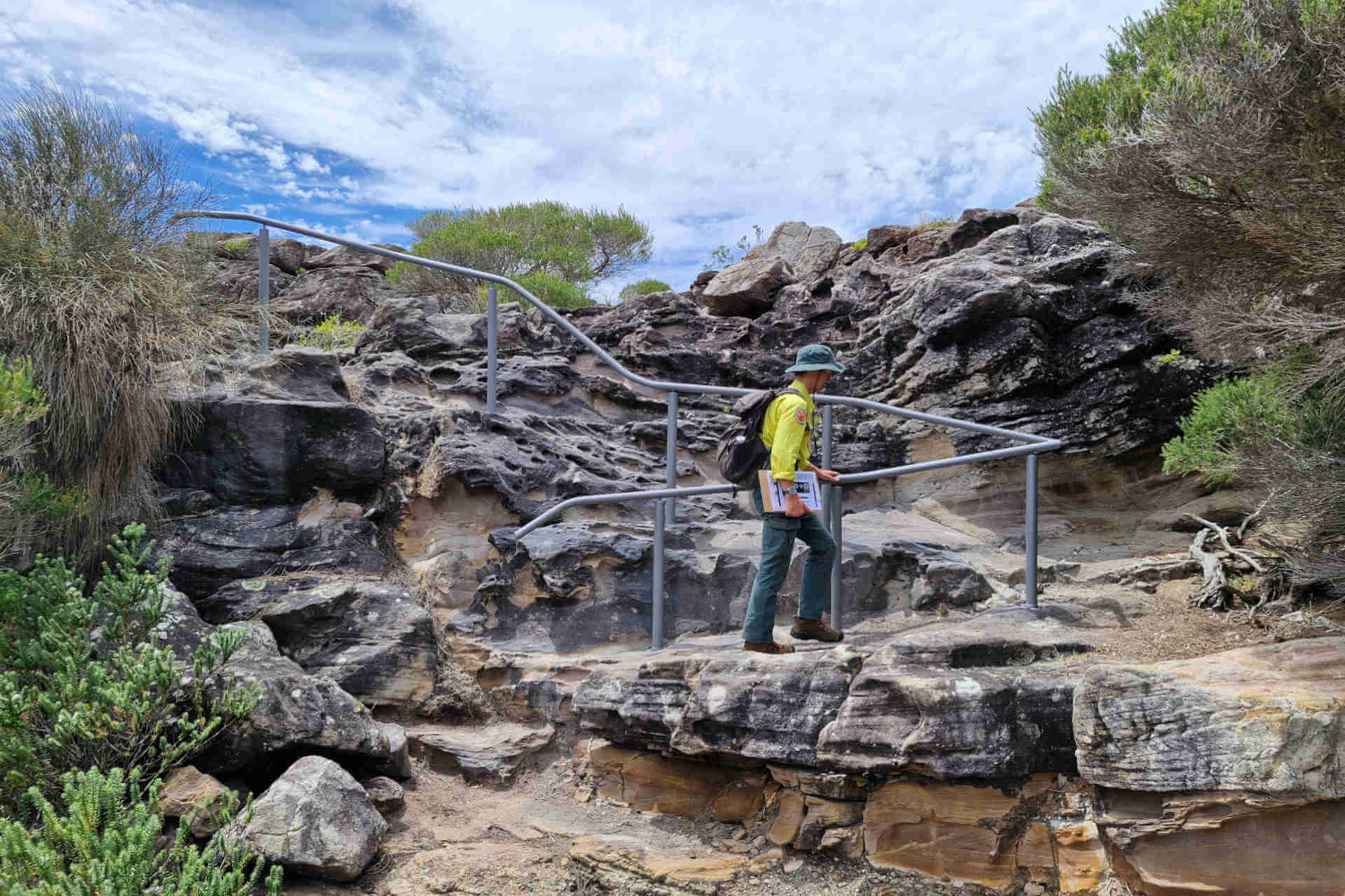 A hiker in full-length sunsafe gear follows a rail up rocky coastal terrain