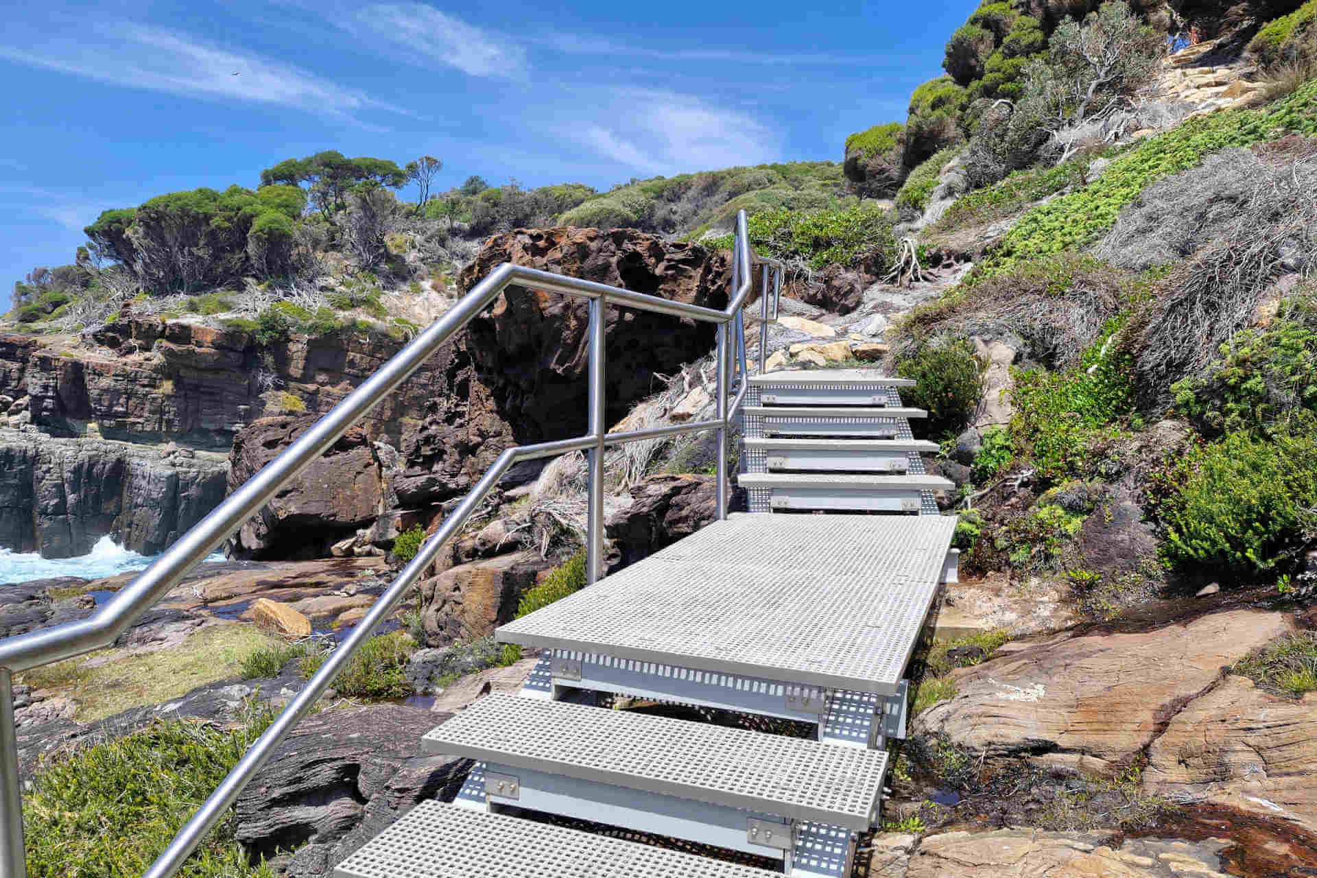 A sturdy-looking metal staircase ascends a rocky seashore, with clusters of windswept cliff-edge trees in the background