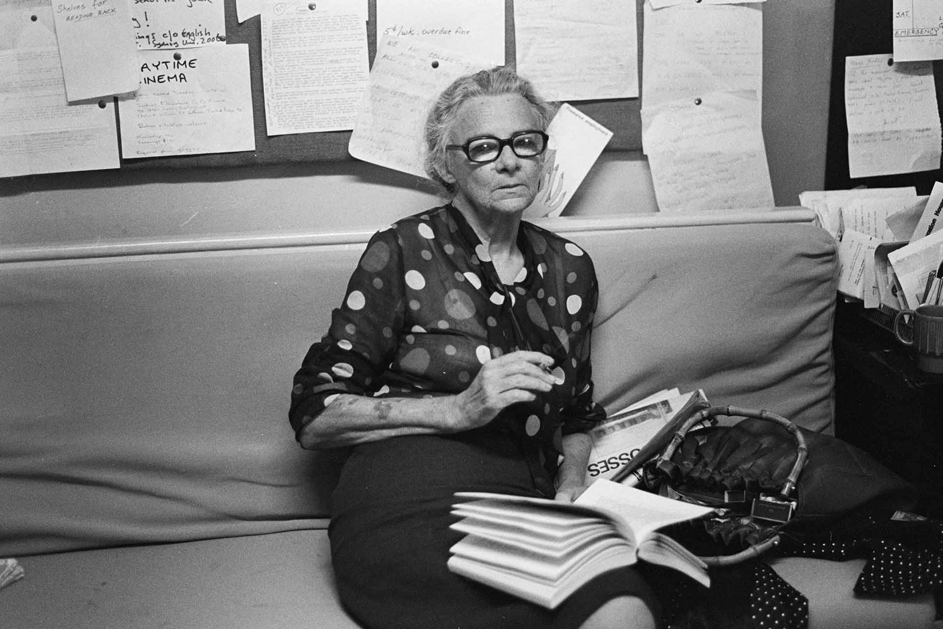 Bessie sitting on a vinyl bench, notice board behind her, handbag beside her, open book on lap and ciagrette in hand.