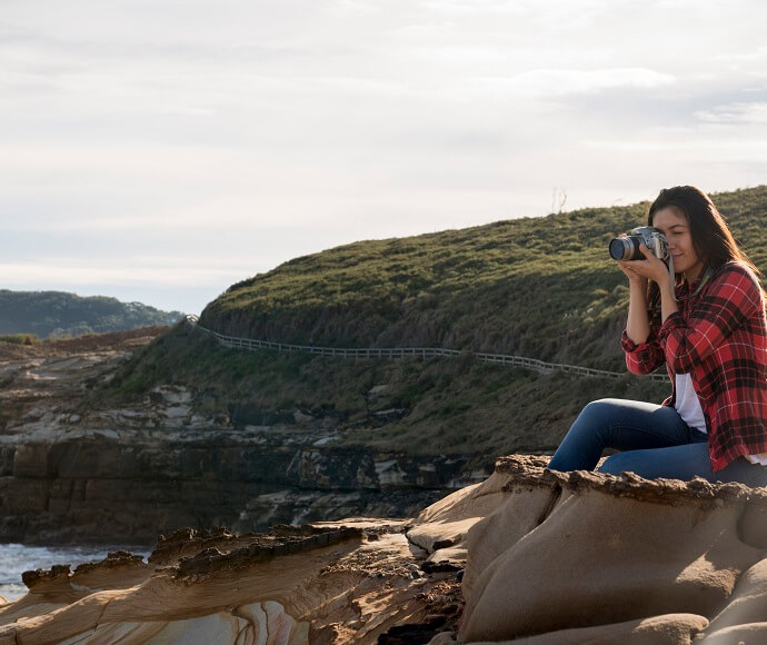 A woman sits on a rocky ledge. She takes a photo of a pretty coastal view. The view includes the Bouddi National Park coastal walk.