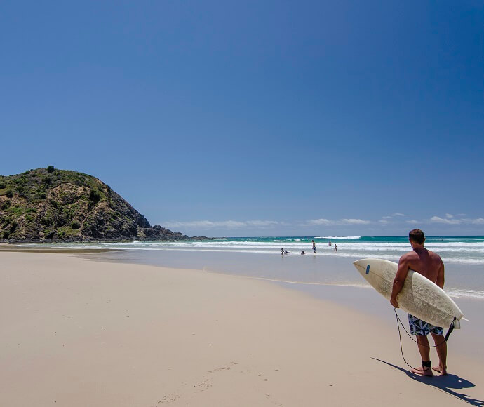A person holding a surfboard looks out towards the ocean on a sunny beach day, with clear blue skies above, waves gently crashing near the shore, and a rocky hill to the left. 