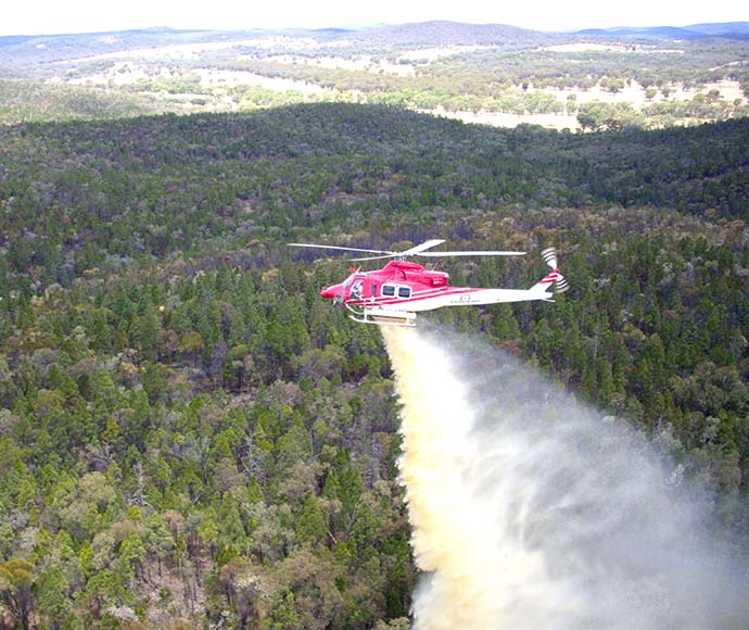 A red helicopter flies over dense bushland, dropping a wave of grey foam down on trees below