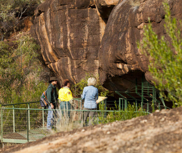Three individuals on a metal viewing platform examining a stratified brown rock formation surrounded by green vegetation.