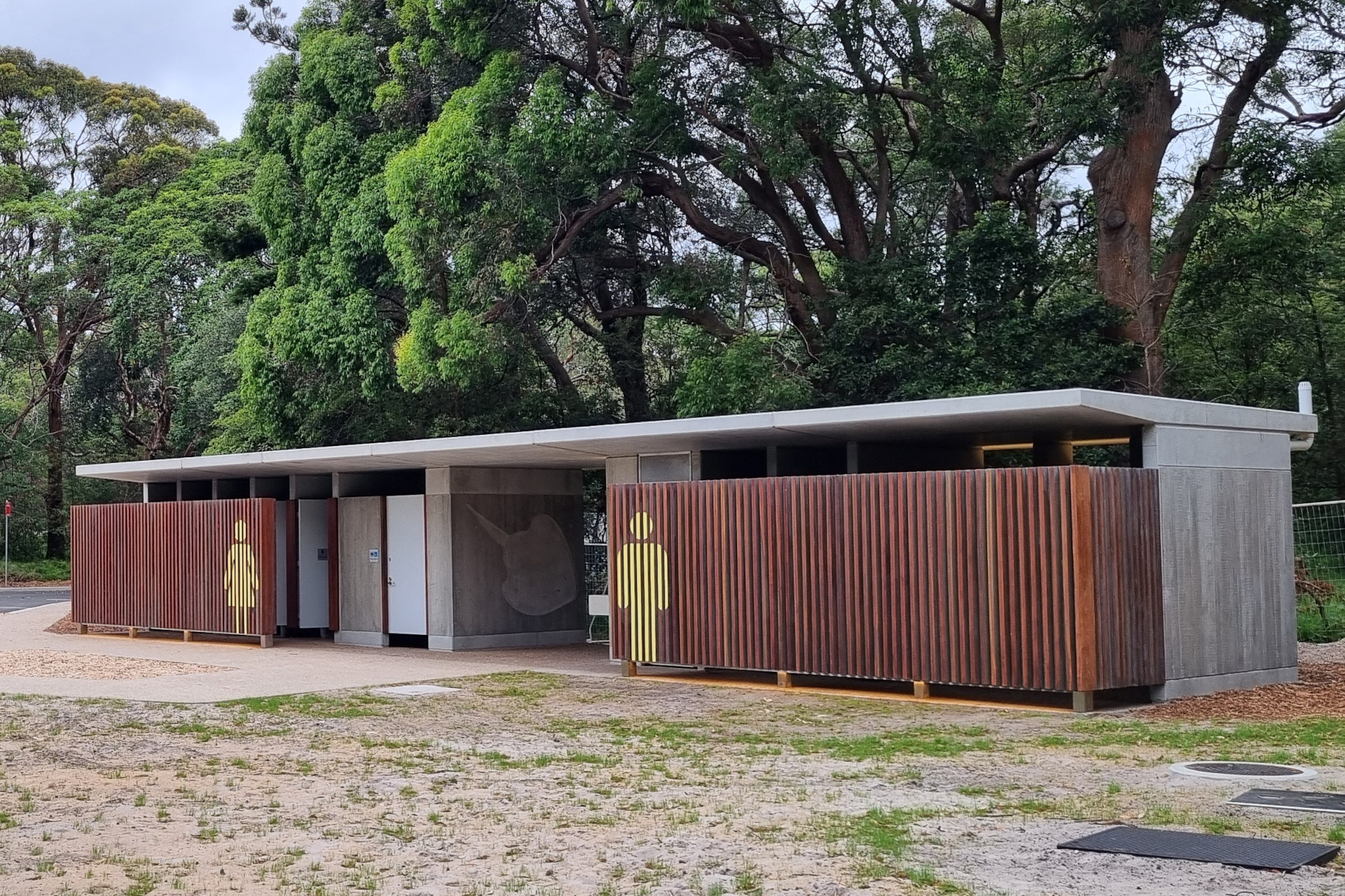 Modern public restroom facility at the Cricket Pitch precinct at Kurnell, Kamay Botany National Park, features separate male and female sections. The structure has a flat roof and is constructed with concrete and wooden slats. The surrounding area is lush with tall green trees.