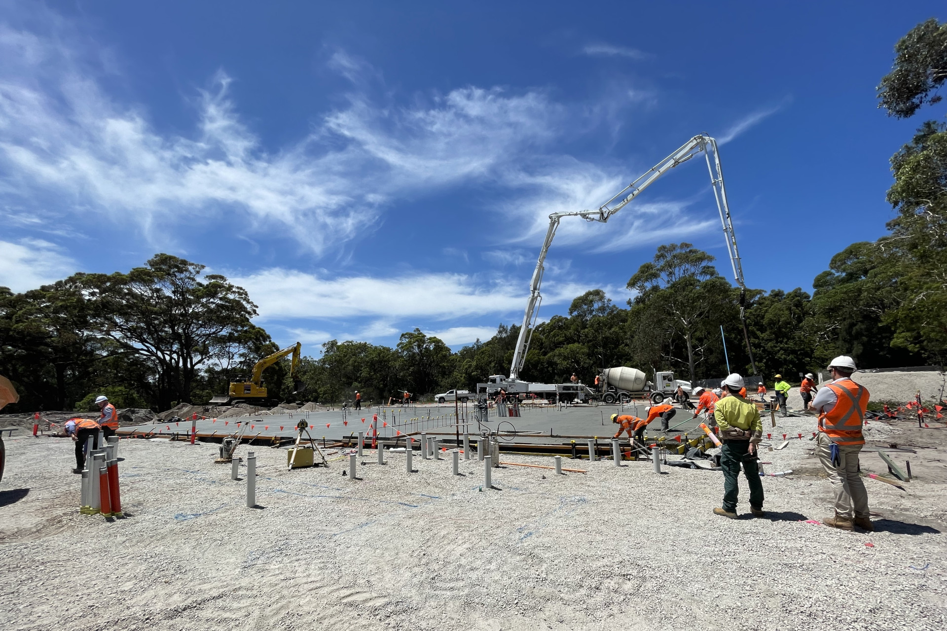 Construction workers in safety gear are working on the Visitor Centre construction site at Kurnell under a clear blue sky. A concrete pump truck is in operation, and various construction equipment and materials are scattered around the site.