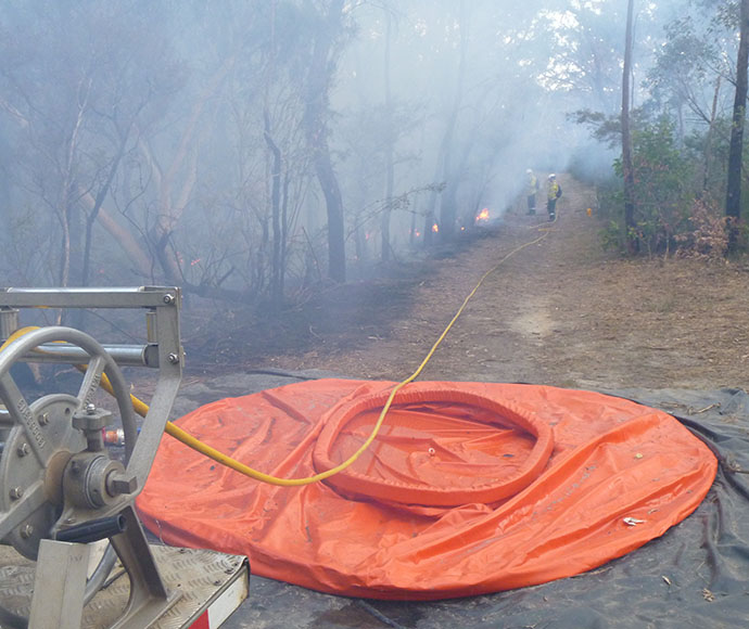 Two firefighters are hosing a line of blackened, charred trees. The bouy wall is bright orange and sits deflated on the ground near the hose reel.