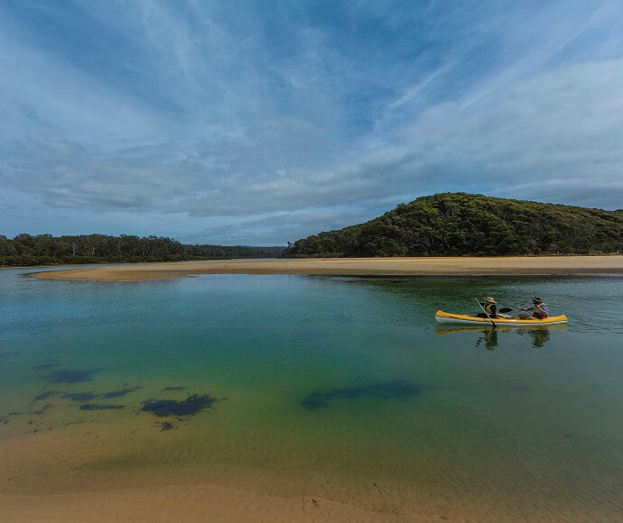 Two people kayaking on a calm body of water with clear visibility to the sandy bottom, near a sandbank, with lush green hills and a blue sky with scattered clouds in the background.