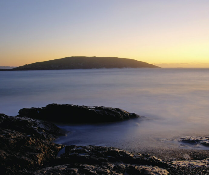 Serene view of Muttonbird Island Nature Reserve at dusk, featuring rocky shores with gentle waves and the island’s silhouette against a gradient sky transitioning from warm yellow to cool blue tones.