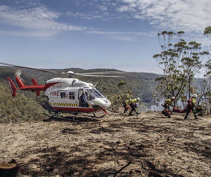 A helicopter has landed on a burnt-out hill, with smoke rising from the valley below. Four firefighters are carrying equipment from the helicopter.