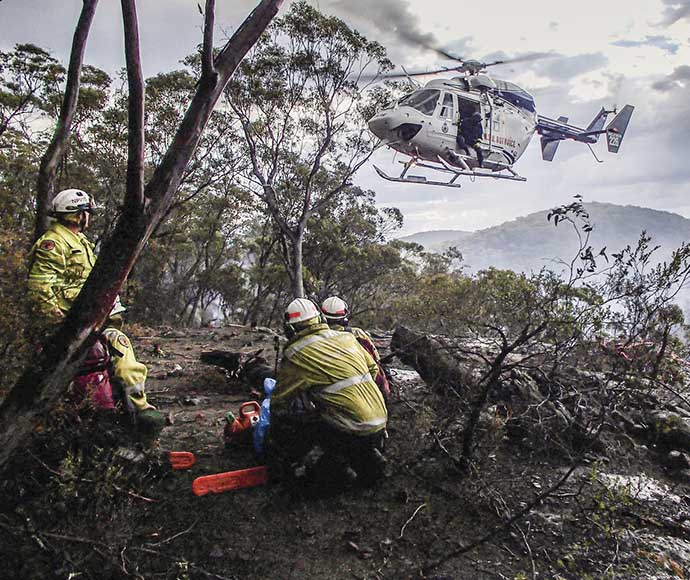 Remote Area Fire Team in uniforms with supplies on a ridge in bushland. A helicopter is hovering above the team.