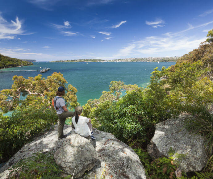 Two people enjoying the view of Sydney Harbour from the Manly scenic walk. They are sitting on a large boulder; the harbour stretches out in front of them with blue skies above.