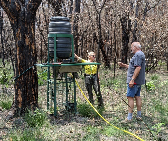 A ranger in uniform stands next to a man in plain clothes while they inspect a drinking station made from a plastic barrel raised above the ground on a metal frame. 