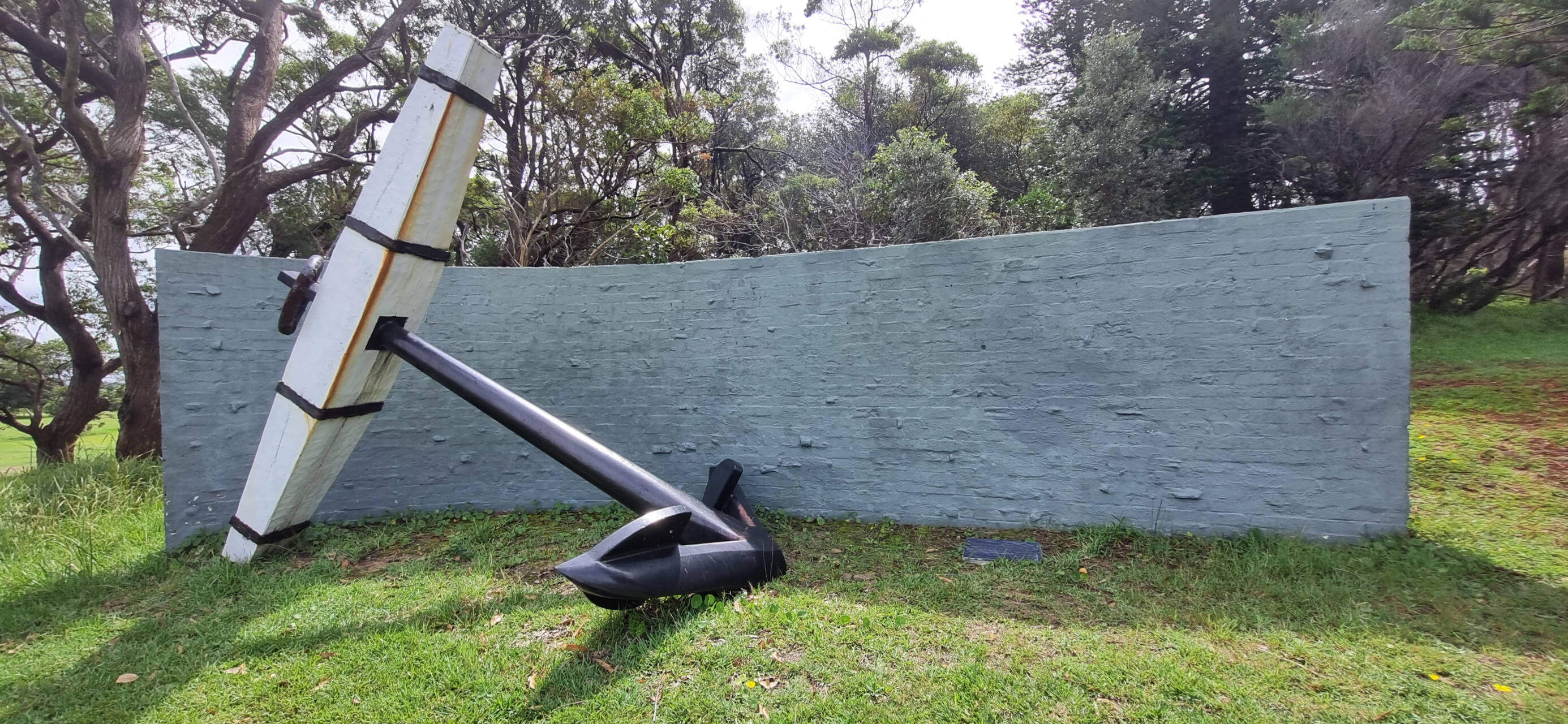 A replica of the HMB Endeavour's anchor is displayed outdoors. It leans against a curved grey stone wall in a grassy area, surrounded by trees.