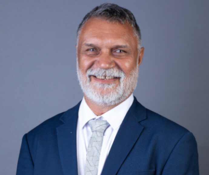 Raymond smiles in a blue suit and silvery-patterned tie that matches his eyes, in front of a deep grey background
