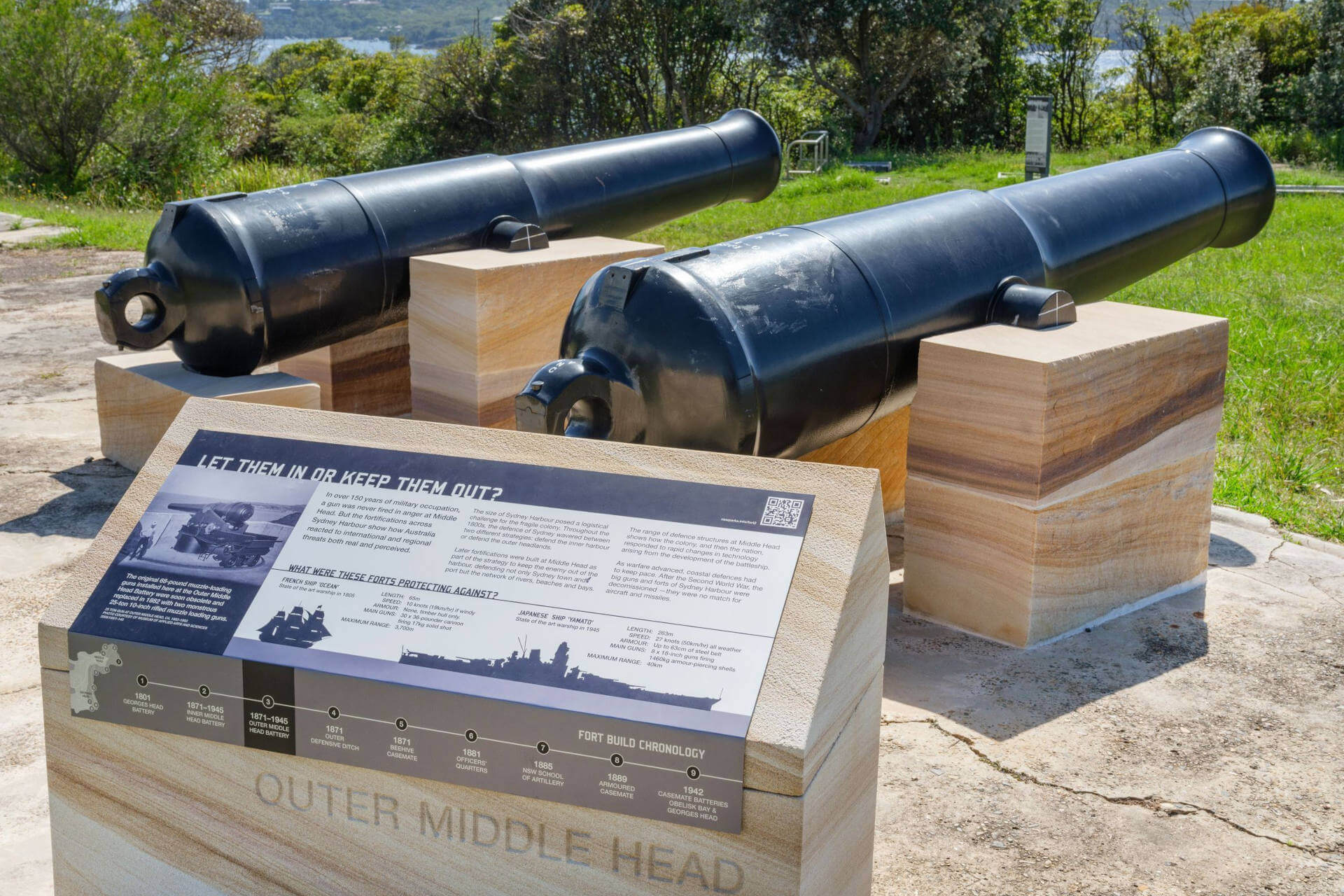 Two large, black restored 68-pounder cannons mounted on stone platforms at Outer Middle Head. In front of the cannons is an informational plaque with text and images explaining the history and significance of the site. The background features greenery and Sydney Harbour.