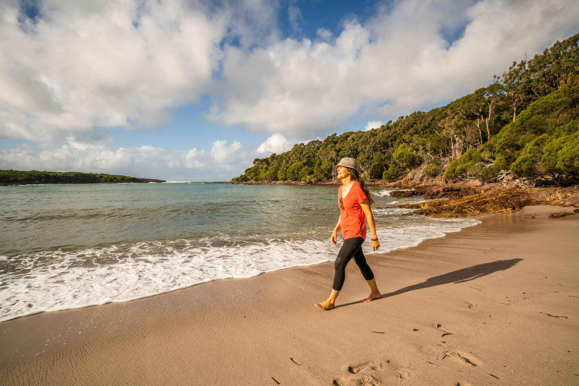 A person walking on a sandy beach beside the ocean, with a backdrop of lush greenery and trees under a partly cloudy sky.