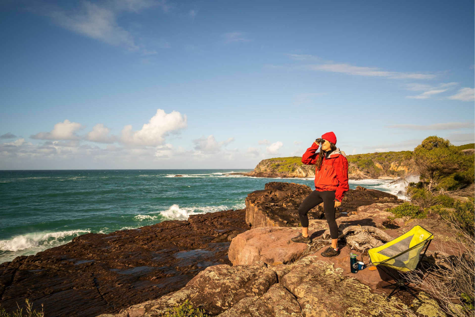 Person in a red jacket and beanie with a pair of binoculars, looking out into the ocean. She is standing on a rocky ledge, the sky is bright blues with a few clouds on the horizon.