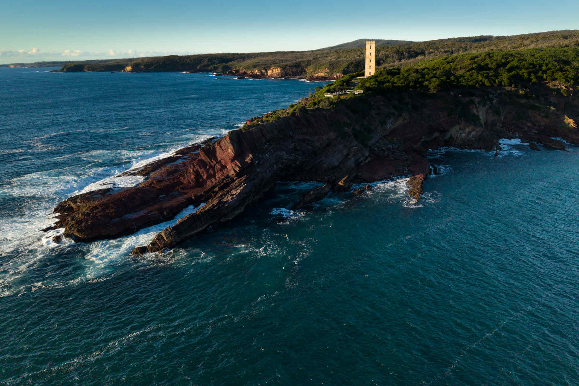 An aerial wide shot of a rocky headland in an azure sea, with a tall old-style brick tower standing out on the headland among the trees
