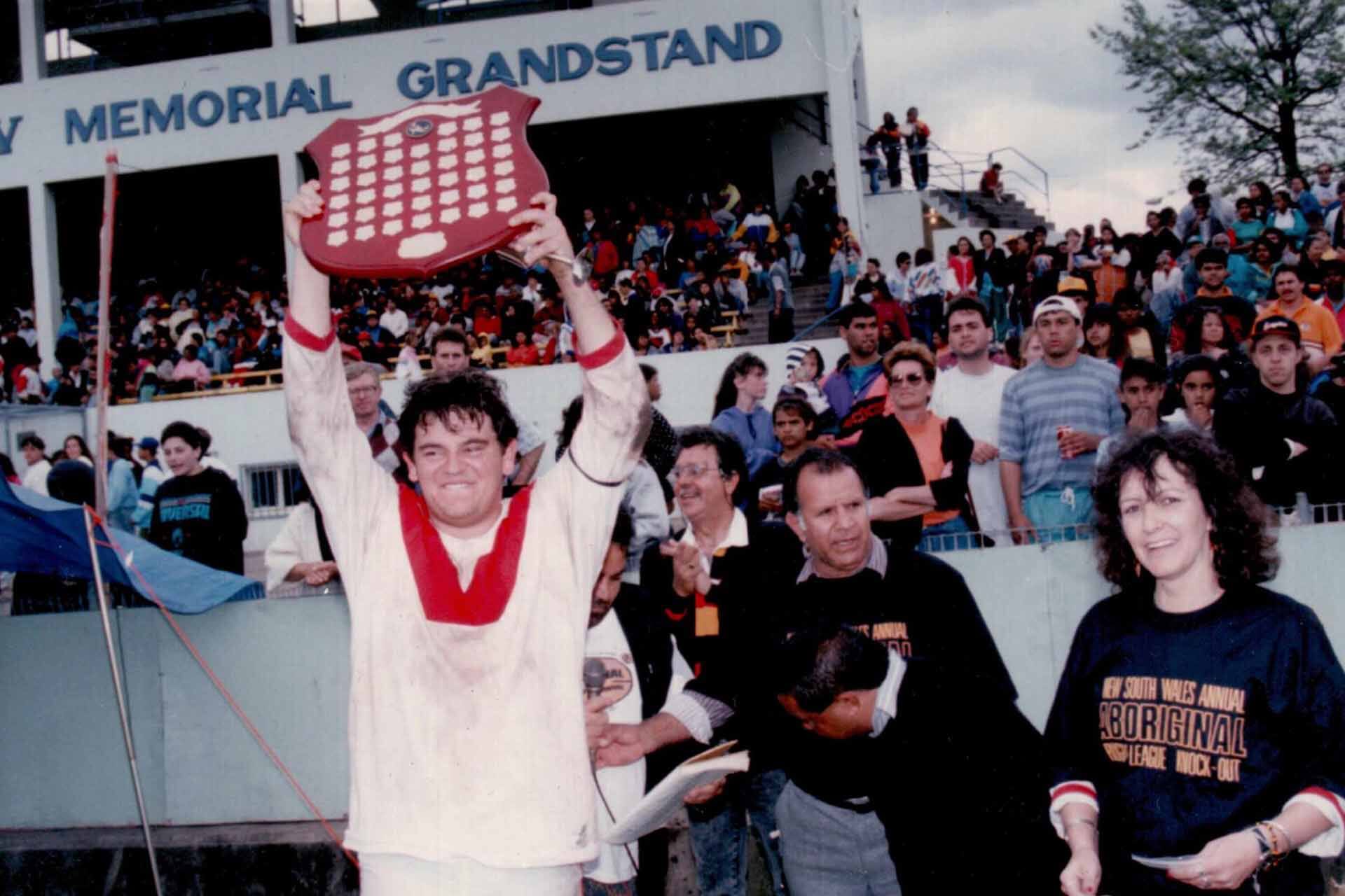 Man in rugby shirt holds trophy aloft while smiling supporters look on