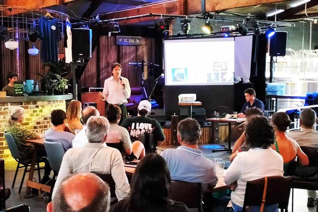 NPWS staff member, holding microphone, standing by screen with prsentation, speaking to an audience gathered around with chairs and tables.
