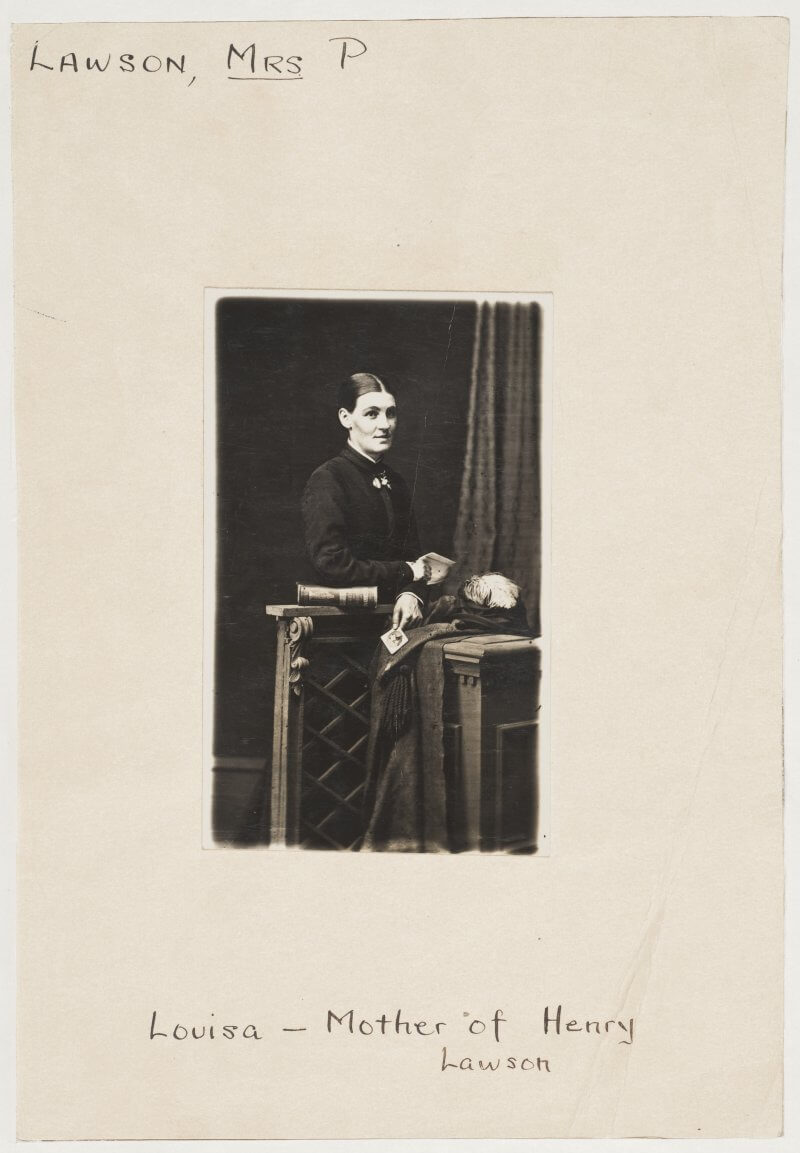 Black and white photo of a woman sitting behind a table, next to a curtain. Her elbow is resting on a book and she is holding a small piece of paper. A hat and coat lie on the table.