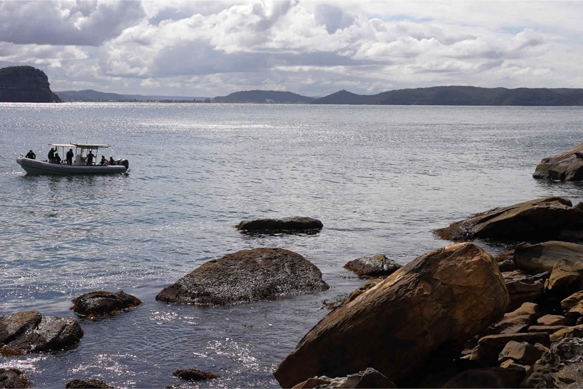 A corner of the coastline comprising boulders and stones; people in a small boat are near the boulders; the sea is flat and calm under a clouded sky