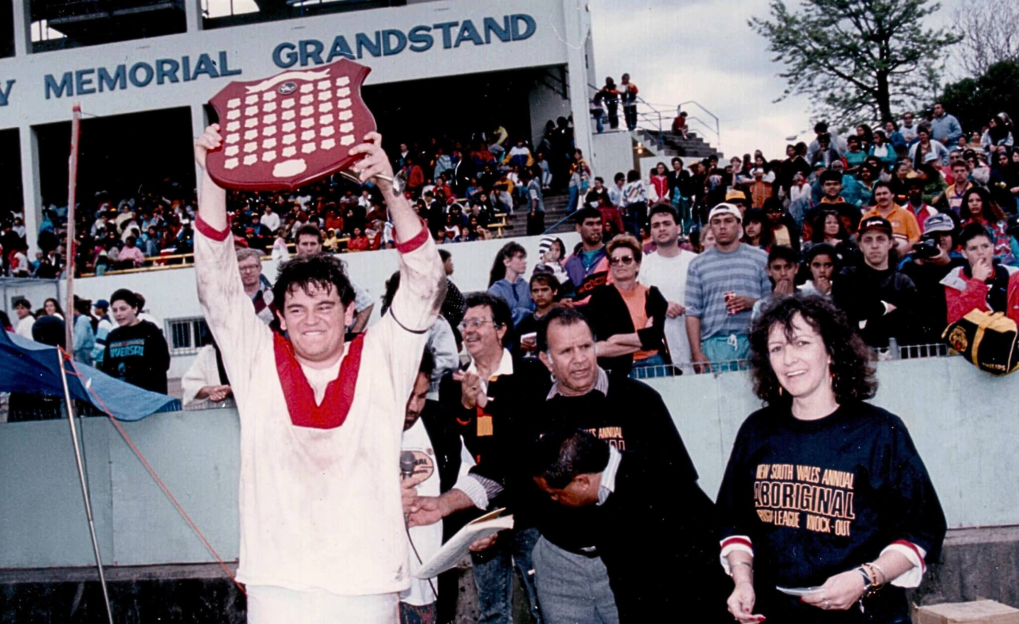 Photo of Brad Cooke at Henson Park, Marrickville, New South Wales, 1992, after winning the under-19s exhibition game. He stands on the field with a proud smile, holding a trophy high above his head. A large crowd of spectators fills the background, capturing a celebratory moment of youth sporting achievement.