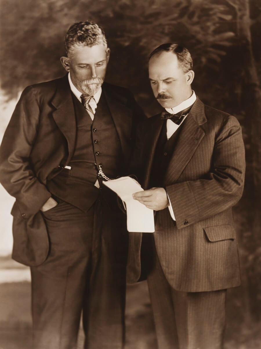 Sepia-toned photo circa 1914 showing Sydney Kidman (left) and Arthur Triggs (right), both dressed in three-piece suits. They are standing close together, looking down intently at a piece of paper held by Arthur Triggs