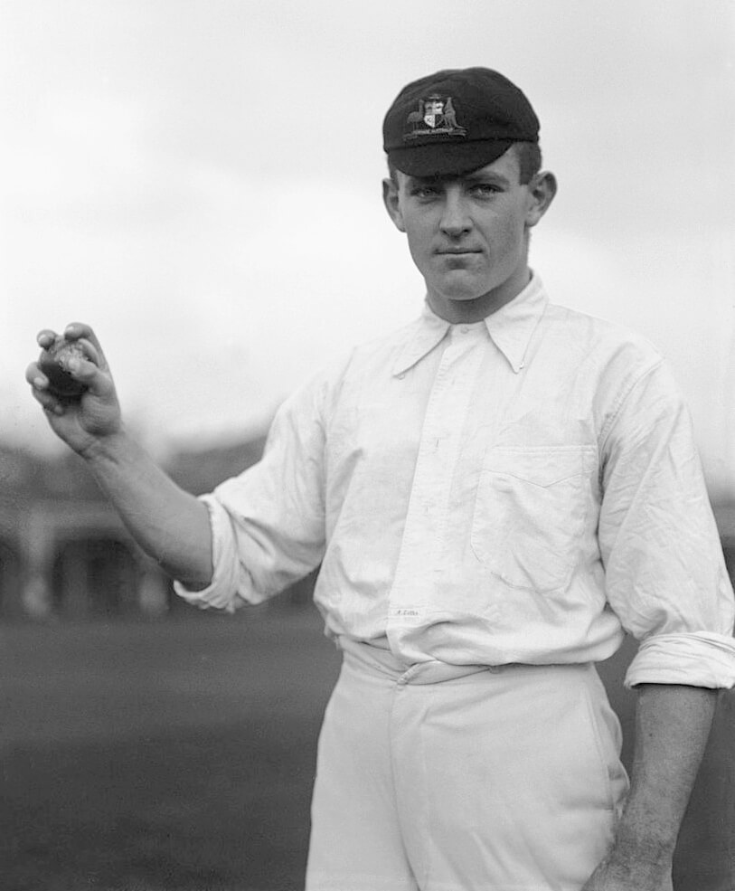 Black and white portrait of Albert Cotter, New South Wales and Australia cricketer, circa 1905. He stands upright, holding a cricket ball up in his right hand. The photo is cropped at the thighs. Cotter wears a dark cap, white long-sleeved shirt, and white pants, looking slightly off-camera with a smile.