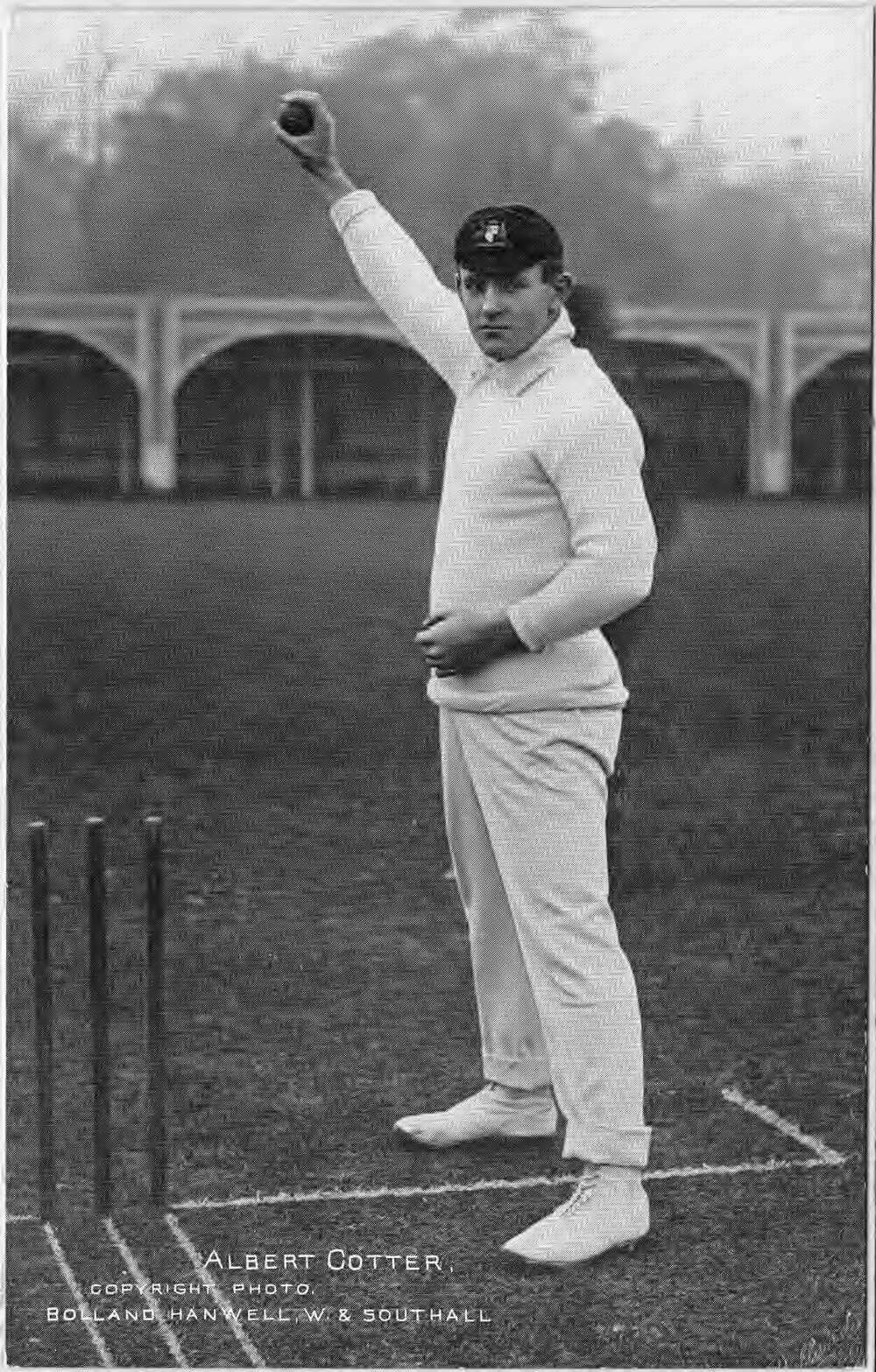 Black and white portrait of Australian fast bowler Albert 'Tibby' Cotter in 1908. He stands upright beside a cricket stand, holding a cricket ball high in his right hand, poised to deliver a ball. Cotter wears a dark cap, white long-sleeved top, white pants, and white shoes.