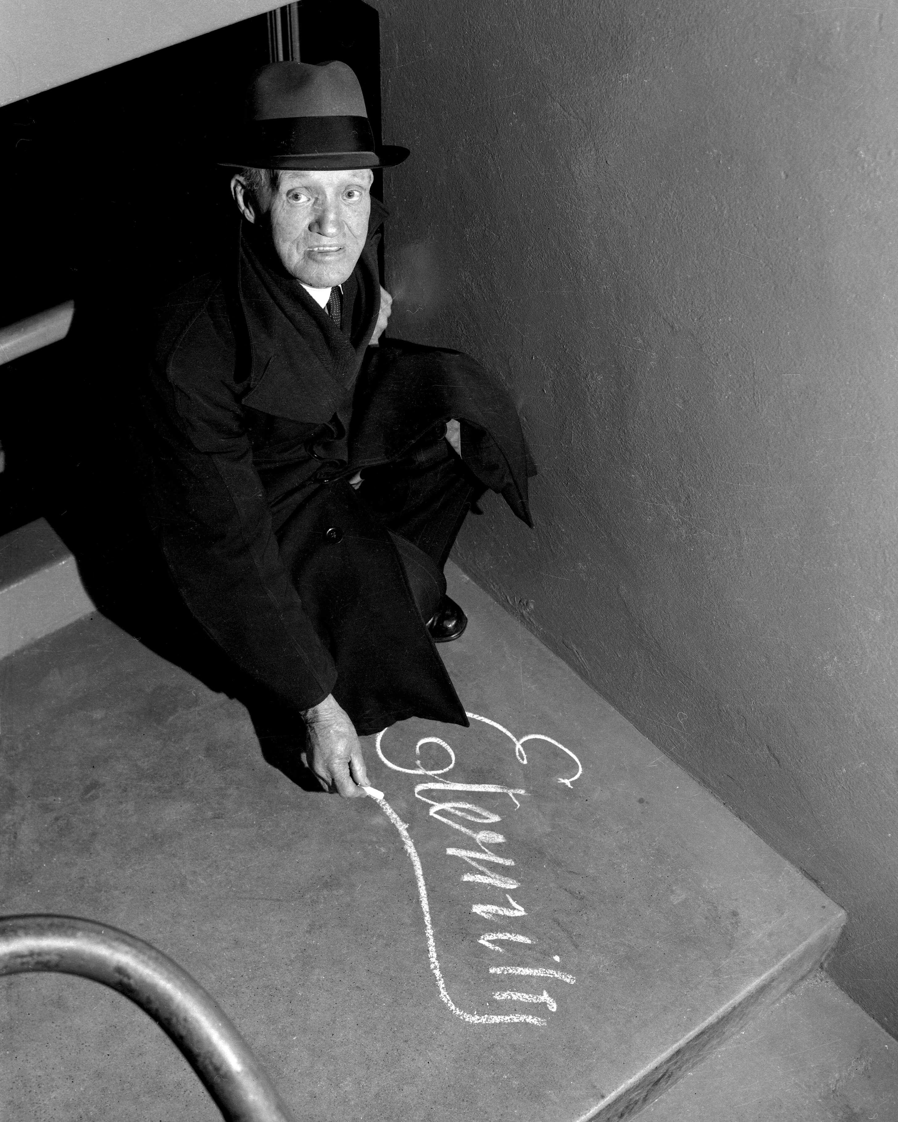 Black and white photograph taken on 3 July 1963, showing Arthur Stace squatting on a Sydney pavement as he writes the word 'Eternity' in chalk. He looks up toward the camera, posing for photographer Trevor Dallen. The word is written in his signature copperplate script.