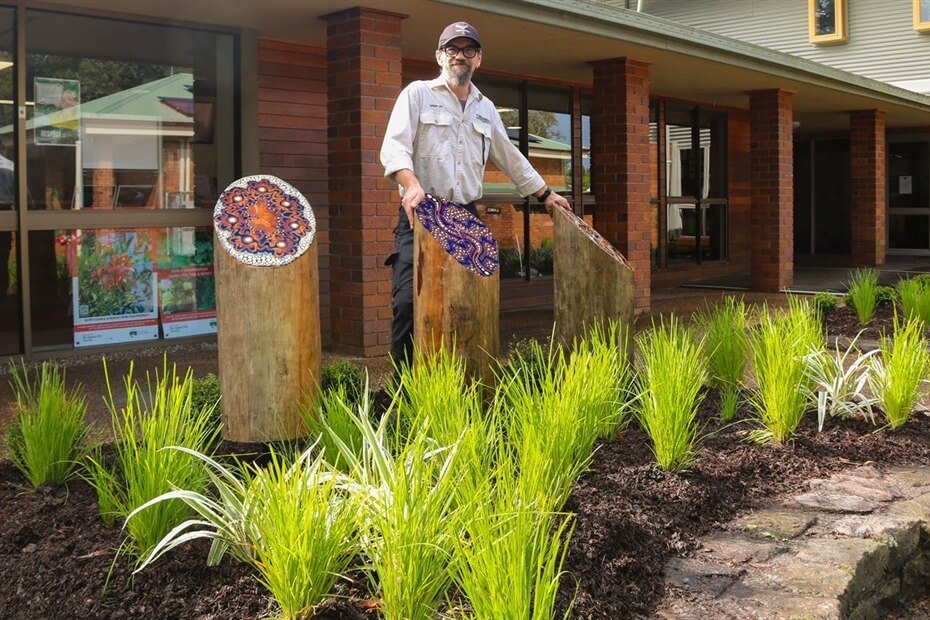 A man stands beside Indigenous artworks on posts in a garden bed with a path and small plants. There is a brick building in the background.