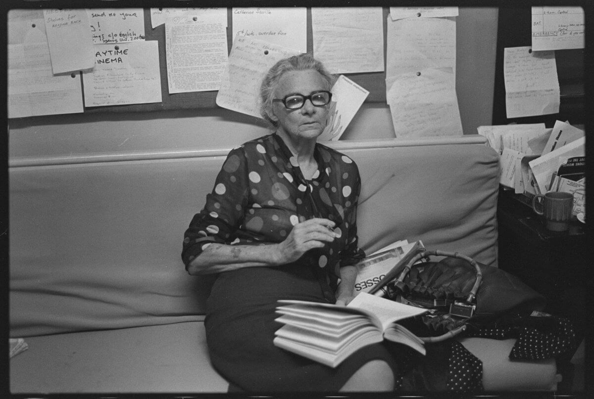 Black and white photo of Bessie Guthrie at Sydney Women’s Liberation House, 1974. She sits on a couch with an open book on her lap, holding a pen in her right hand. She has short hair, wears black-rimmed glasses, and a polka dot chiffon shirt with rolled-up sleeves. Her handbag and jacket are beside her. Behind her is a board covered in handwritten papers. A small side table holds stacked papers and a cup.