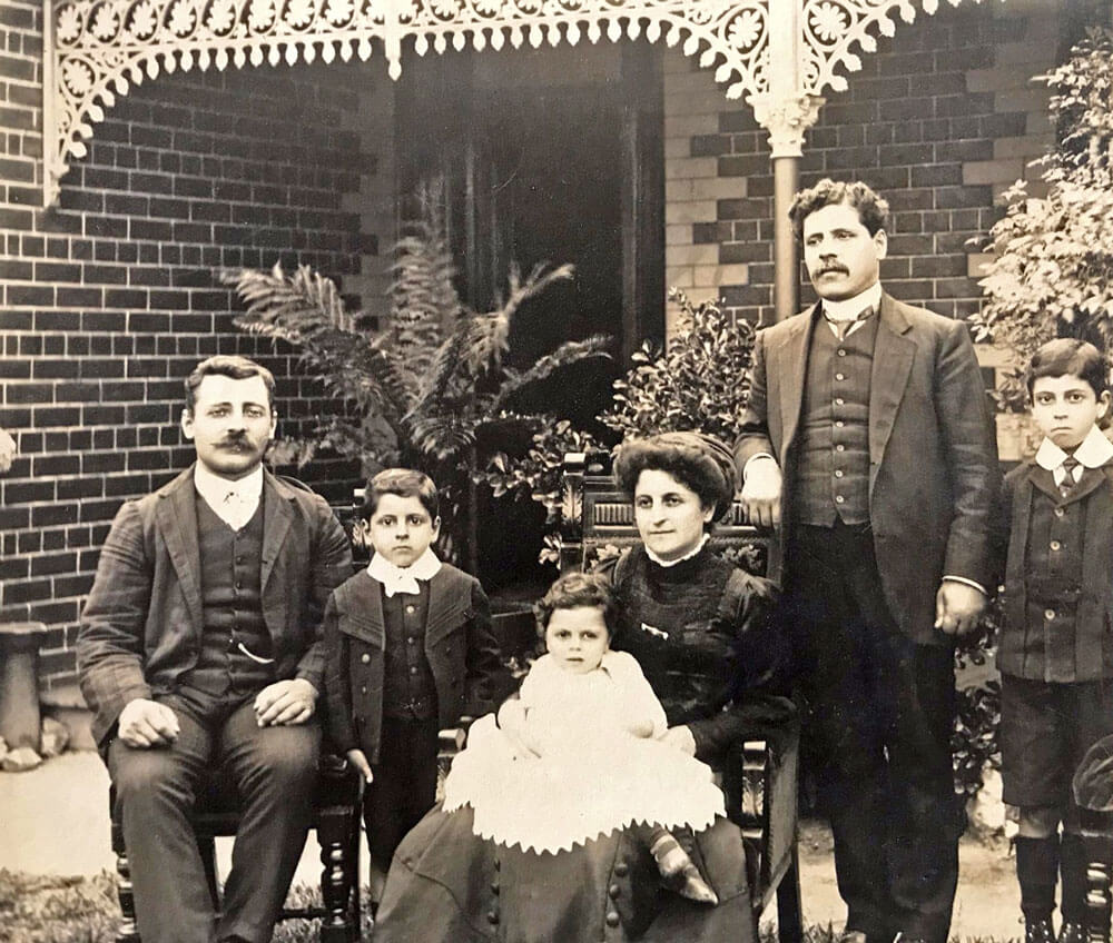 Sepia photo of Betro Abicare and family, circa 1911, taken in the front yard of a brick house. From left to right: a man believed to be Betro’s brother Nicholas is seated, followed by Victor, baby Frederick held by seated Julia, Betro standing, and Claude. The group is arranged in a relaxed family pose, with a mix of seated and standing figures.