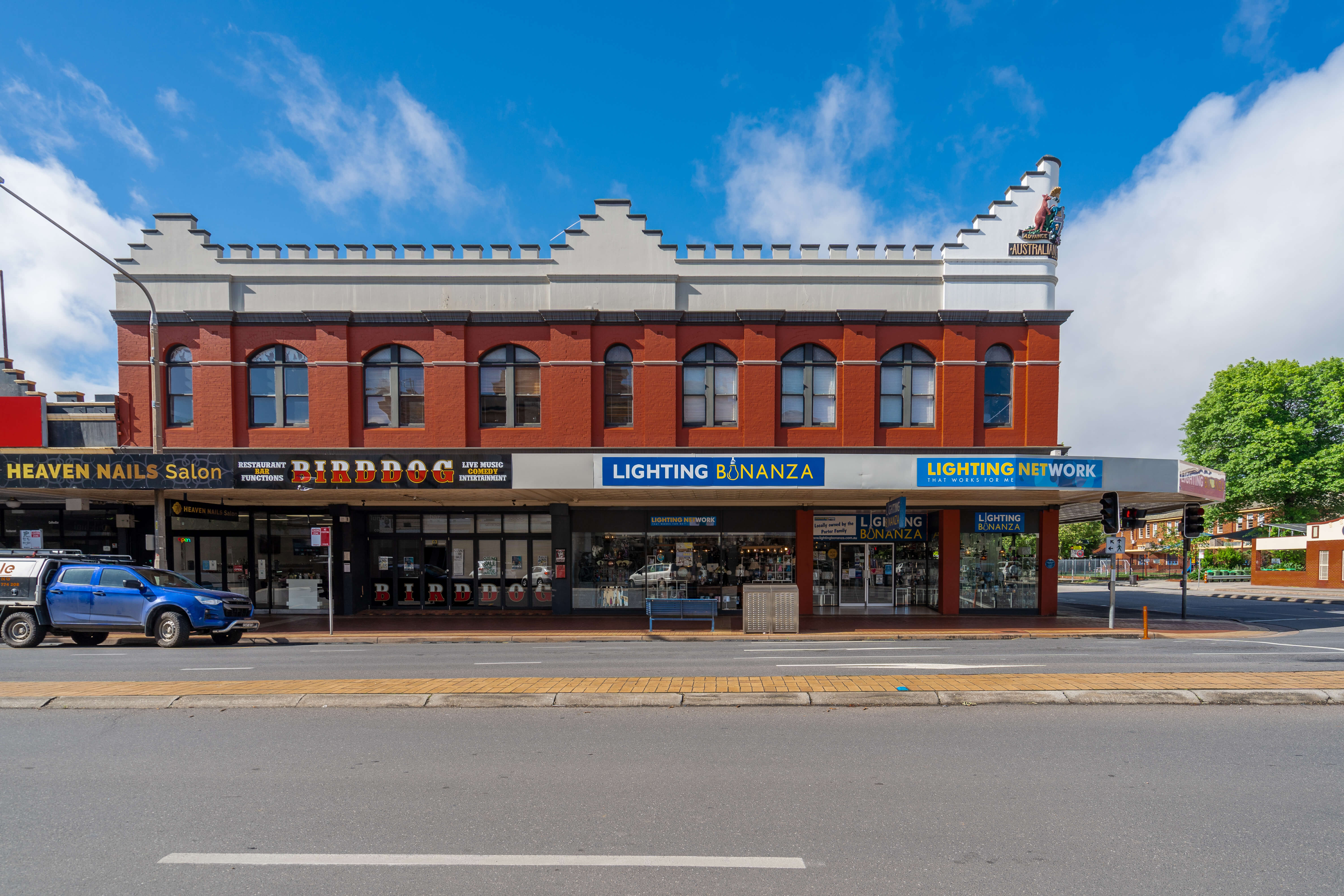 Photo of the current building on the former site of The Big Store in Albury, taken from across the road. The two-storey building has a brown brick upper level and a creamy white roof. The ground floor is now divided into several businesses, including 'Heaven Nails Salon,' 'Birddog Restaurant,' and 'Lighting Bonanza.'