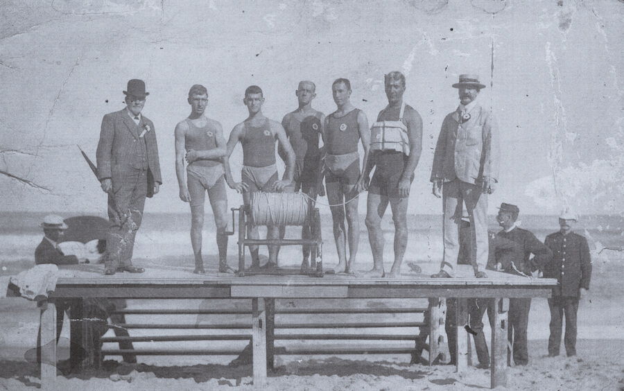 Black and white group photo of the Bondi Surf Bathers Life Saving Club demonstration team at Newcastle Beach in 1908. The group consists of seven men; the two on the far left and right wear suits and hats, while the five men in the center wear singlets and early-style swimwear briefs or trunks.