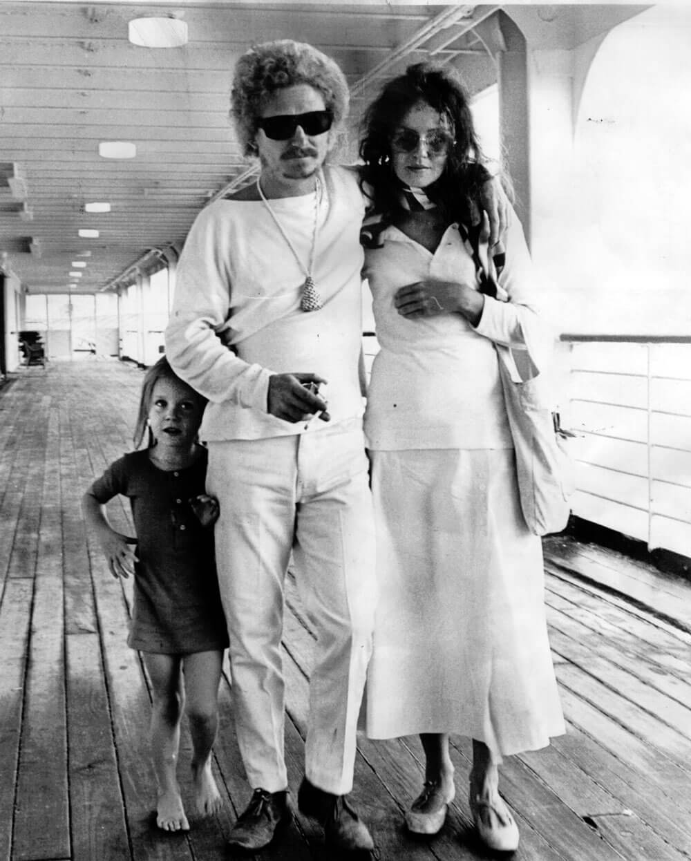Black and white photo of Australian artist Brett Whiteley arriving back in Sydney on 2 November 1969 with wife Wendy and 4.5-year-old daughter Arkie. Brett stands in the center wearing a white long-sleeved top, sunglasses, and a long necklace with a pendant. Wendy is on the right, dressed in a long-sleeved white top, long white dress, sunglasses, and carrying a white handbag. Arkie stands barefoot on the left, at Brett’s waist height, wearing a dark long-sleeved dress.
