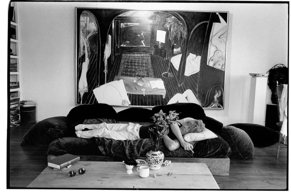 Black and white photo of Brett Whiteley at Lavender Bay in 1976, asleep on a couch with cushions. He is wearing a short-sleeved dark top, long white pants, and dark socks. Behind him is a large painting on the wall. In front of the couch, a coffee table holds a vase of flowers, a cup and saucer, and several books.