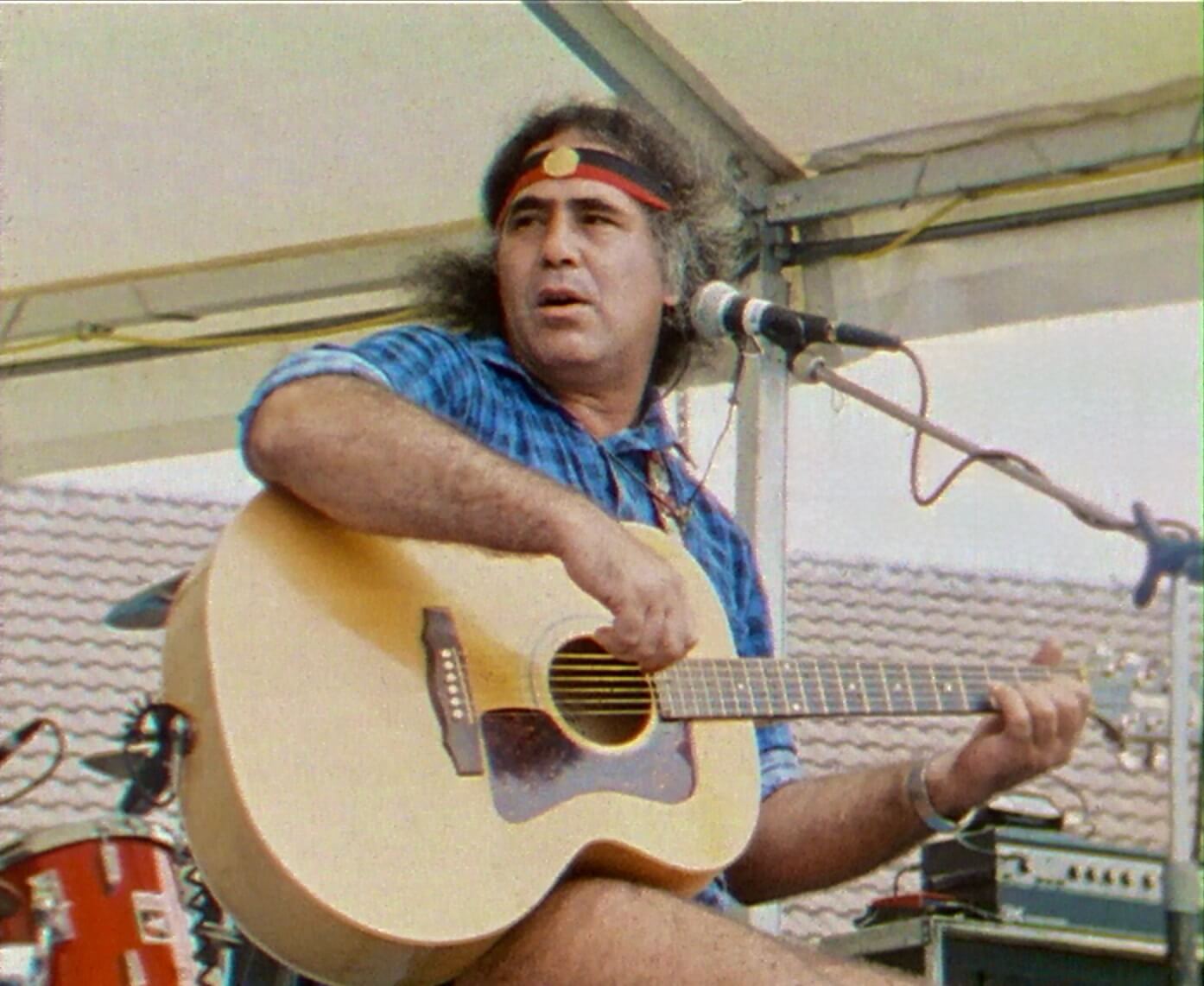 Kev Carmody performing at the Building Bridges Concert at Bondi Pavilion in 1989. He is seated, holding a guitar, with a microphone stand in front of him. He wears a blue checked shirt and a headband featuring the pattern of the Aboriginal flag.