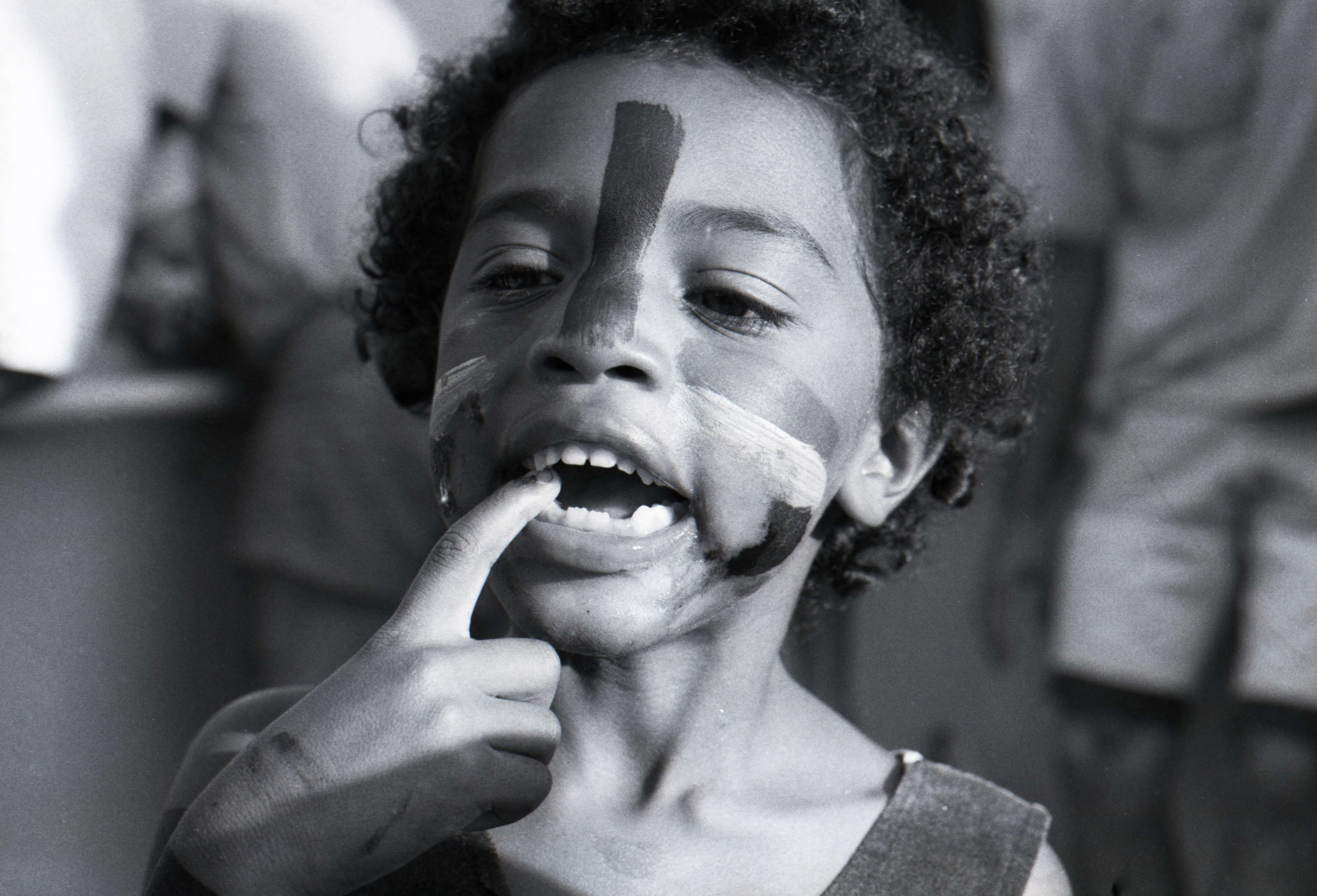 Black and white photo of a young child attending the 1989 Building Bridges Concert. The child has face paint and is captured in a candid moment, biting their second finger.