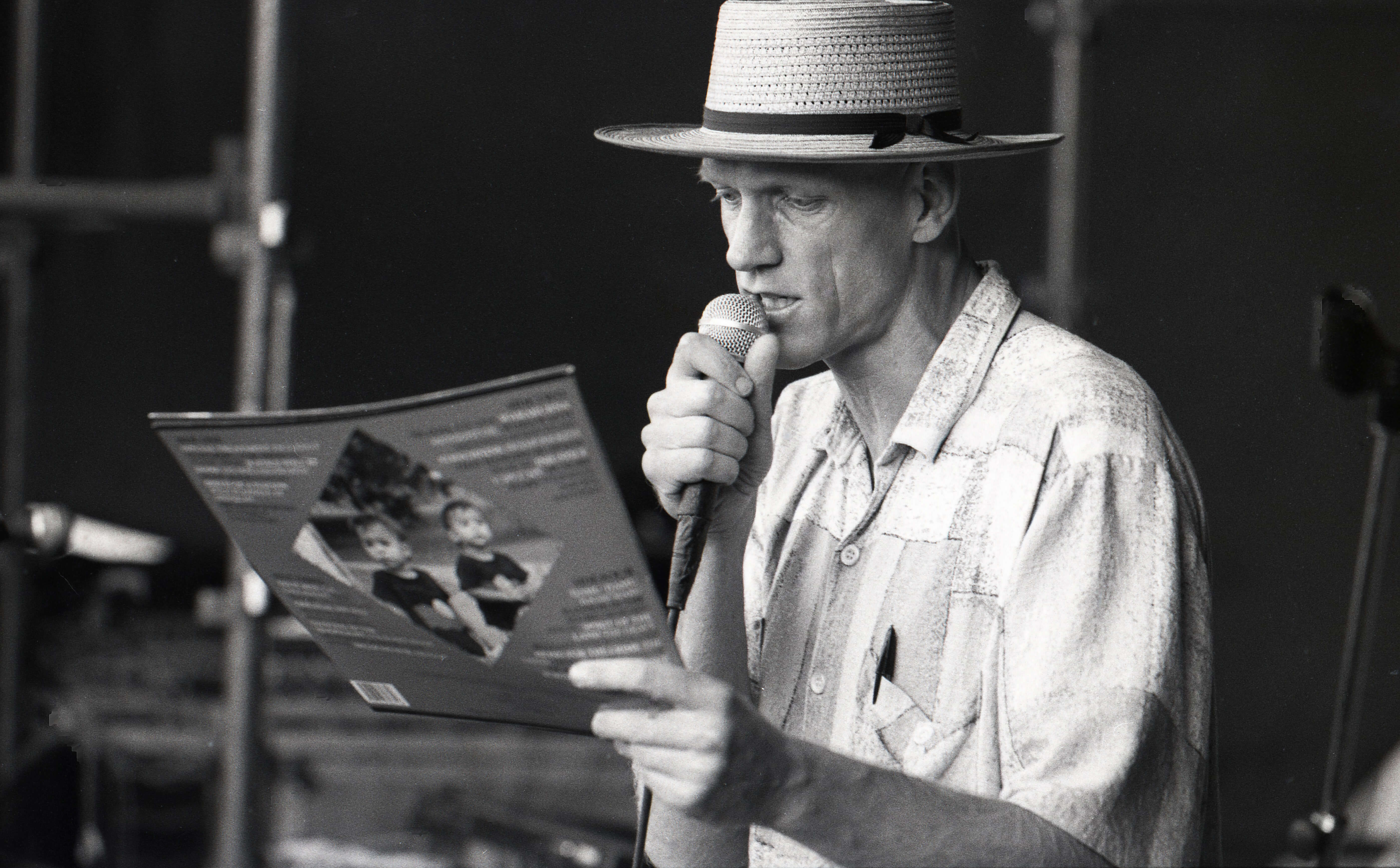 Black and white photo of Peter Garrett MCing at the 1989 Building Bridges Concert. He is wearing a hat and a short-sleeved shirt, holding a microphone in one hand and reading from a brochure in the other.