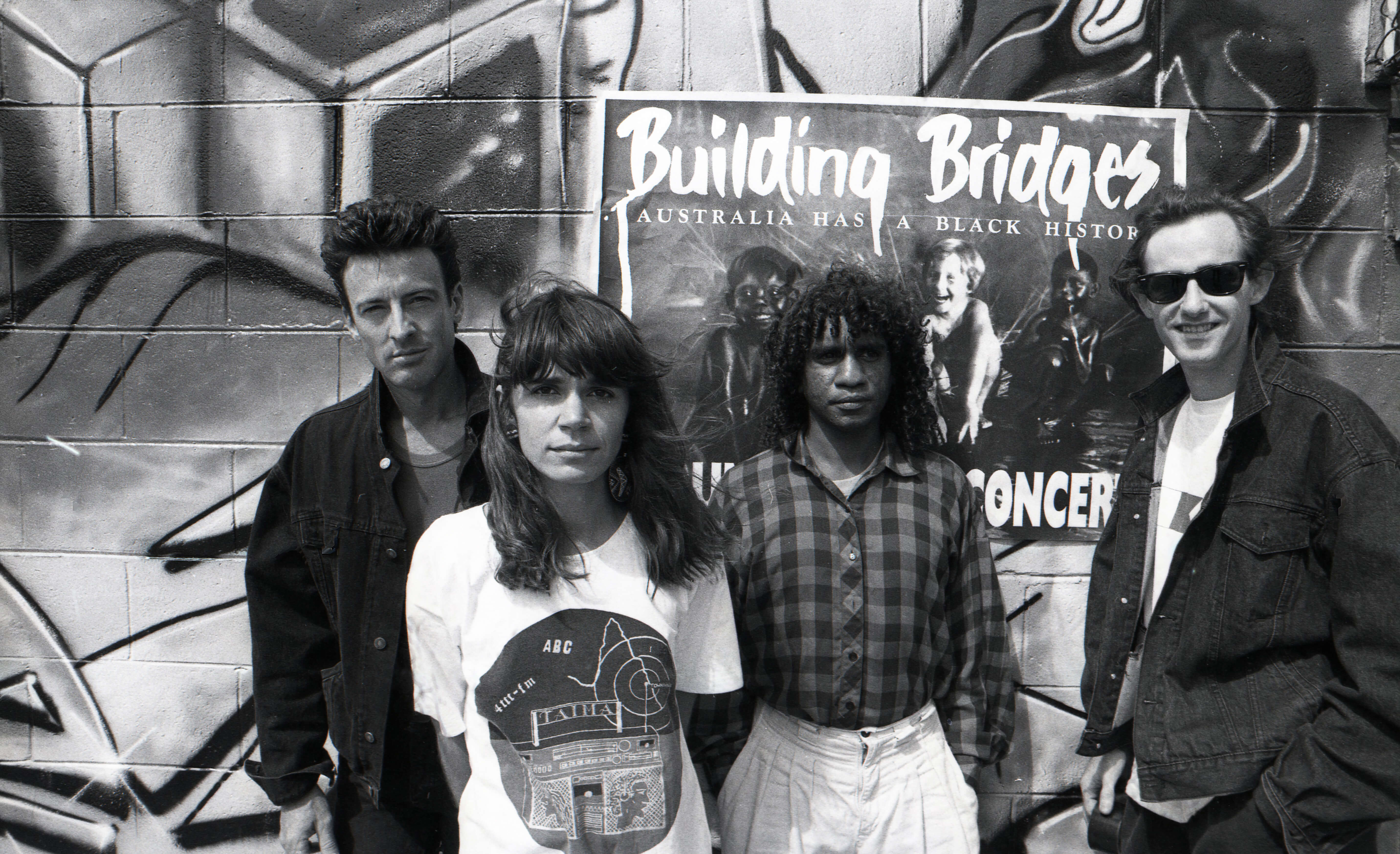 Black and white promotional photo from 1989 featuring Rob Hirst, Sharon Carpenter, Bart Willoughby, and Mark Callaghan standing in front of a graffiti-covered wall. A poster for the Building Bridges Concert is pasted on the wall behind them. The group poses for a photoshoot promoting the Building Bridges Concert album, capturing a gritty, urban aesthetic reflective of the era and the movement.