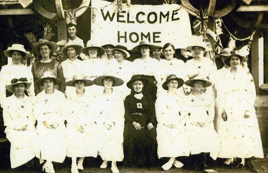 Black and white group photograph taken around 1919 in Macarthur Park, showing 20 women of the Camden Red Cross Sewing Circle. The women are dressed in long white dresses and hats, with the front row seated and the back row standing. Behind them hangs a large 'Welcome Home' banner with Union Jack flags on poles. Mrs. Honor Harris (née Sidman), wearing black, stands out among the group.