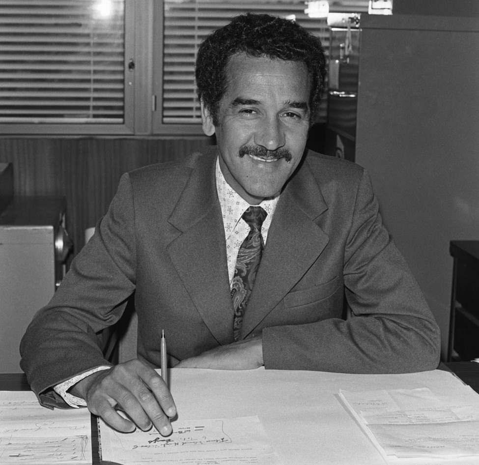 A black-and-white photograph of Indigenous activist and leader Charles Perkins behind his desk at the Department of Aboriginal Affairs in Canberra, taken in 1974. Perkins is smiling with a pen in hand and work papers neatly set out. Photographer unknown.