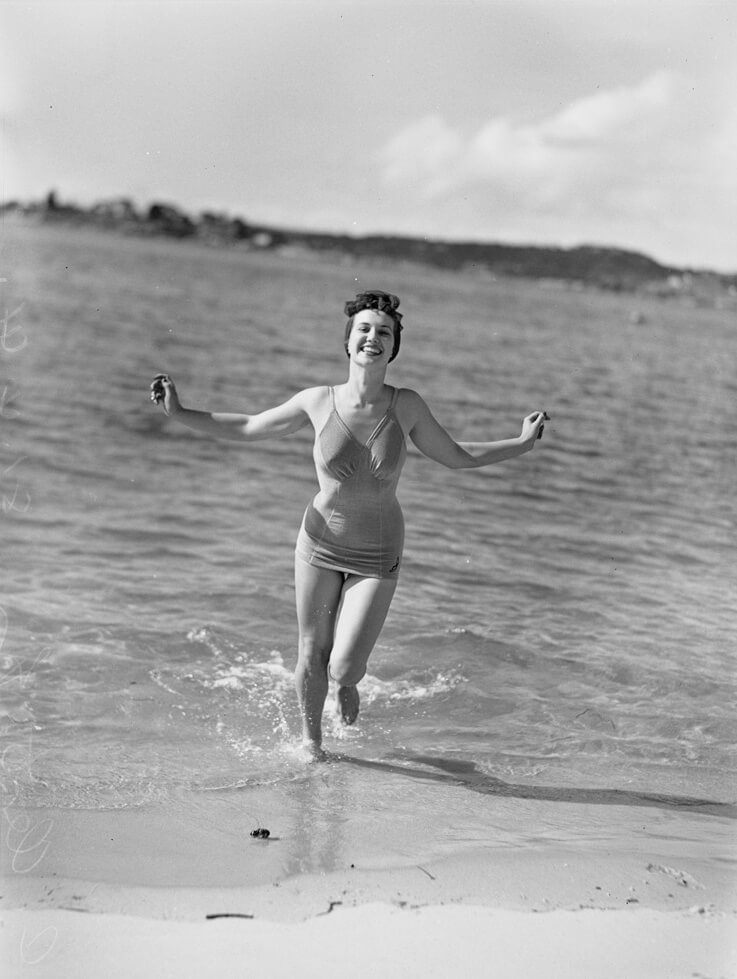 Black and white photo of Charmian Clift modeling a swimsuit for PIX Magazine, dated 21 April 1941. She is running from the ocean toward the beach, wearing a one-piece swimsuit. Her hair is tied up, arms stretched out joyfully, and she is smiling widely with her teeth showing, capturing a moment of carefree energy and happiness.