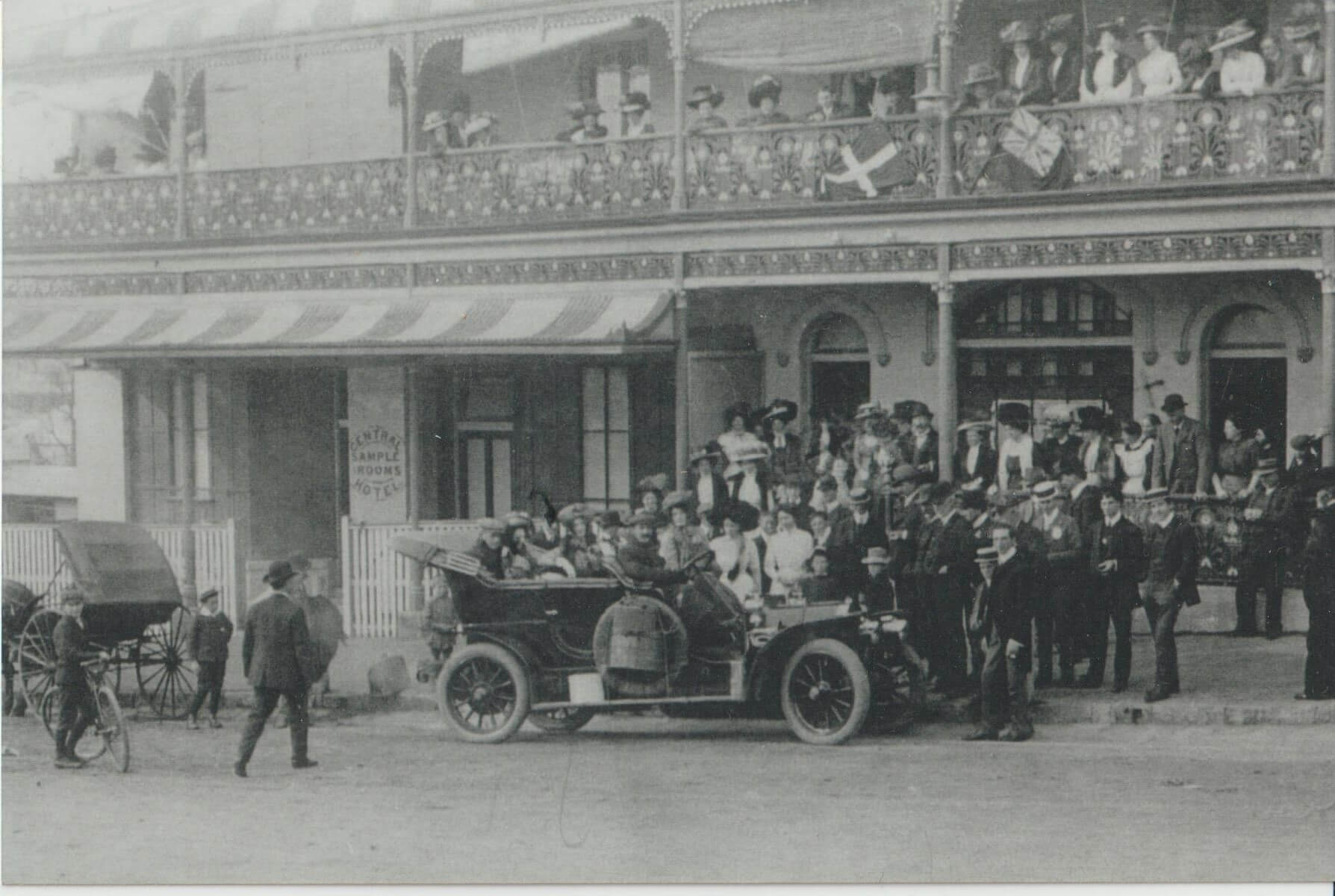 Black and white image of men and women in an old motor car. The car is on the street next to a building which is a hotel, with people looking out from the bottom floor entrance and on the balcony above.