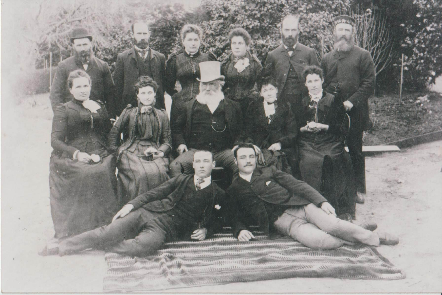 Black and white image of 7 men and 7 women. The men are wearing suits, with three of them wearing hats and the women are wearing Edwardian dresses. Two of the men are lying down in front of the others who are sitting and standing behind them. The family is standing outside with bushes and trees behind them.