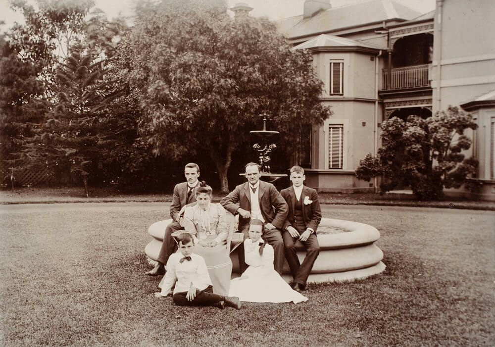 Black and white image of Dorothea and her family sitting on and around a fountain outside. Her father and brothers are all wearing suits while her mother and herself are wearing long dresses. There is a large patch of grass around the fountain with trees and part of a building in the background.